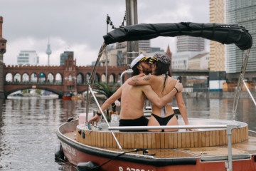 A couple enjoying a private Romantic Special Hot Tub Boat experience on the river in Berlin, relaxing in warm water as they cruise past the Oberbaumbrücke at sunset. The floating bathtub is softly decorated for romance, creating an intimate and unforgetta