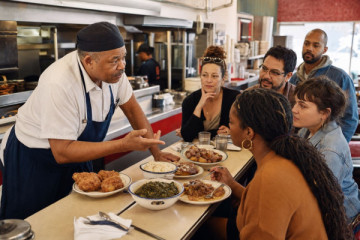 A local cook explaining generous regional dishes to guests gathered around a neighbourhood lunch counter