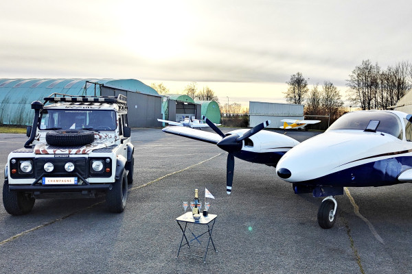 The photo is taken on the airfield tarmac and there is the Defender on the left and the twin engine Seneca III on the right. A mini table with glasses and a bottle of champagne is in the center.