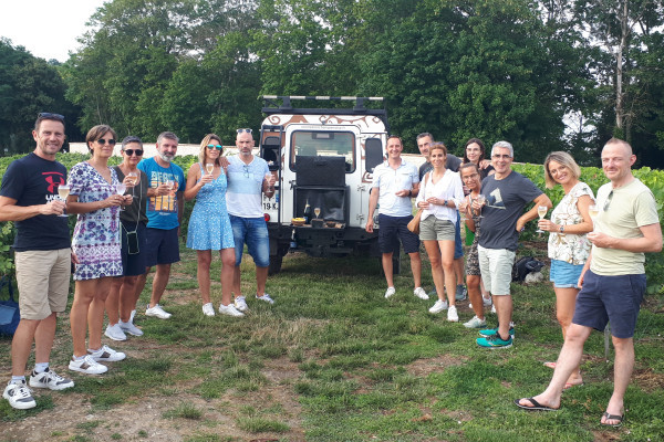 the photo is on a vineyard road, the Defender is in the background and 14 people are on either side of the back of the vehicle a glass of champagne in hand.