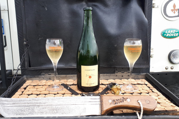the bar of the vehicle is open, a bottle of champagne slashed in the center on each side two glasses of champagne full with the name Connexion Champenoise and in the foreground the sabre.
