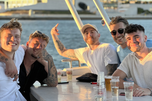 a group of 5 young, handsome men sitting by the table, drinking beer, smiling and having fun on a boat party