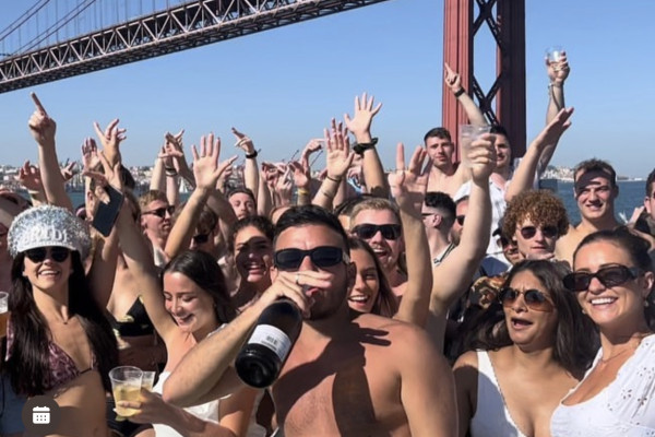 a big group of young people on a boat dancing, drinking, waving their hands and smiling for the photo