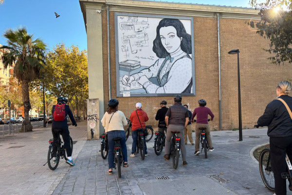 Cyclists riding toward a large black-and-white portrait mural dedicated Hedy Lamar on a sunny day in Valencia
